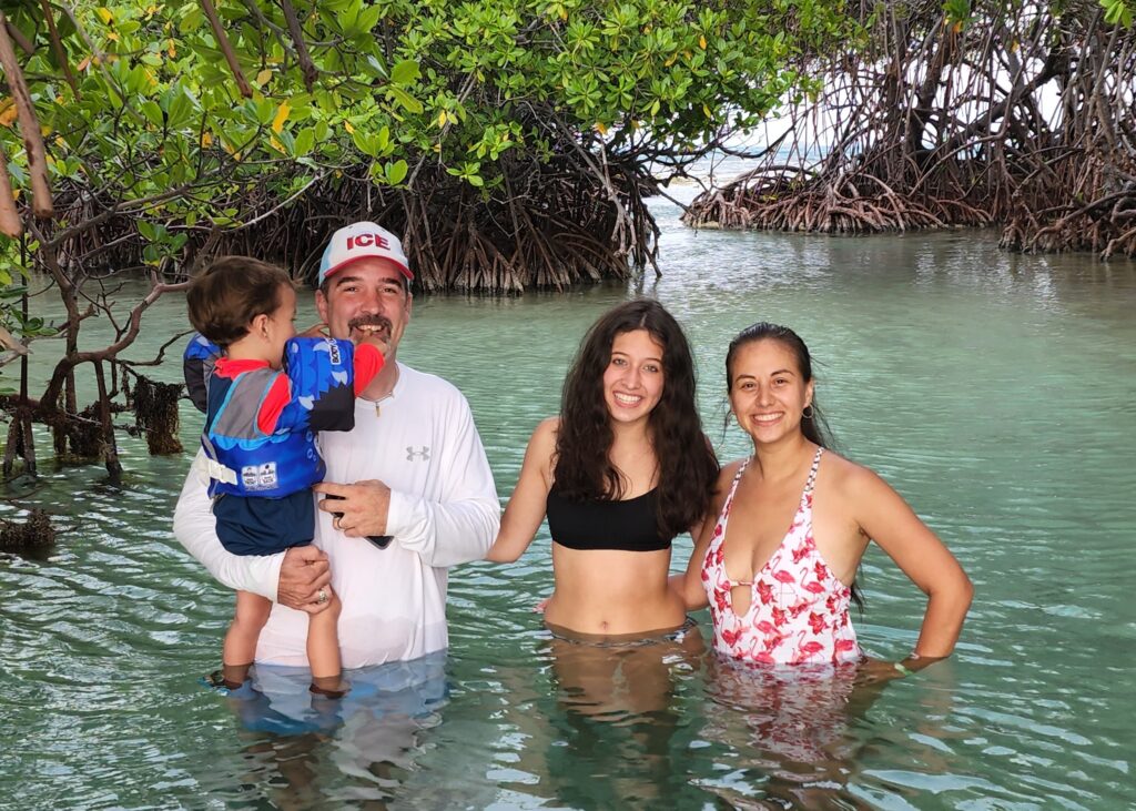 A family, including young children, smiles while wading in the pristine waters of La Parguera's Enrique Cay. Surrounded by lush mangroves, this safe and serene experience is part of Bucketlist Tours’ Mystic Waters Under the Sun & Stars, offering a perfect blend of natural beauty and relaxation for all ages.