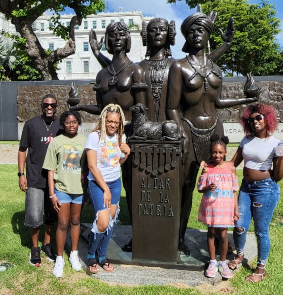 Family traveling to Puerto Rico experiencing Bucketlist Tours' Old San Juan Ageless Elegance, Heritage and Spirits private tour, posing by the iconic 'Altar de la Patria' , monument to the Puertorrican Women, surrounded by lush greenery and cultural heritage.