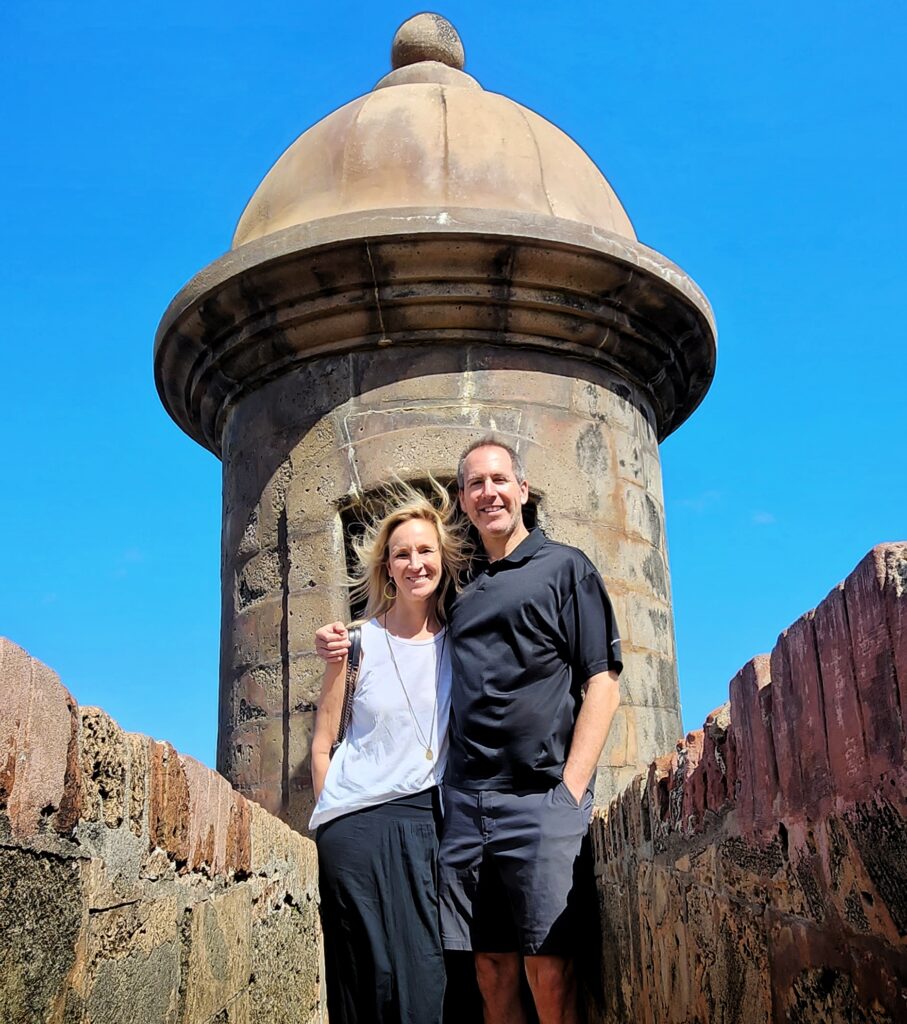 Couple experiencing to Puerto Rico enjoying Bucketlist Tours’ Old San Juan Ageless Elegance, Heritage, and Spirits private tour, exploring the historic walls of Old San Juan, standing by a guard tower with breathtaking views of the ocean under a clear blue sky.