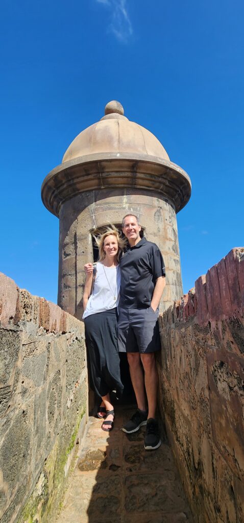 Couple experiencing to Puerto Rico enjoying Bucketlist Tours’ Old San Juan Ageless Elegance, Heritage, and Spirits private tour, exploring the historic walls of Old San Juan, standing by a guard tower with breathtaking views of the ocean under a clear blue sky.