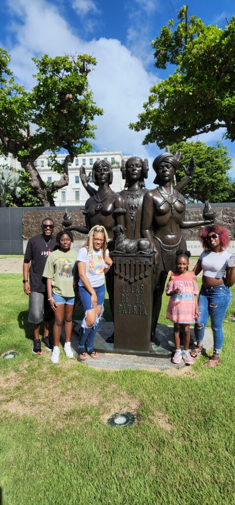 Family traveling to Puerto Rico experiencing Bucketlist Tours' Old San Juan Ageless Elegance, Heritage and Spirits private tour, posing by the iconic 'Altar de la Patria' , monument to the Puertorrican Women, surrounded by lush greenery and cultural heritage.