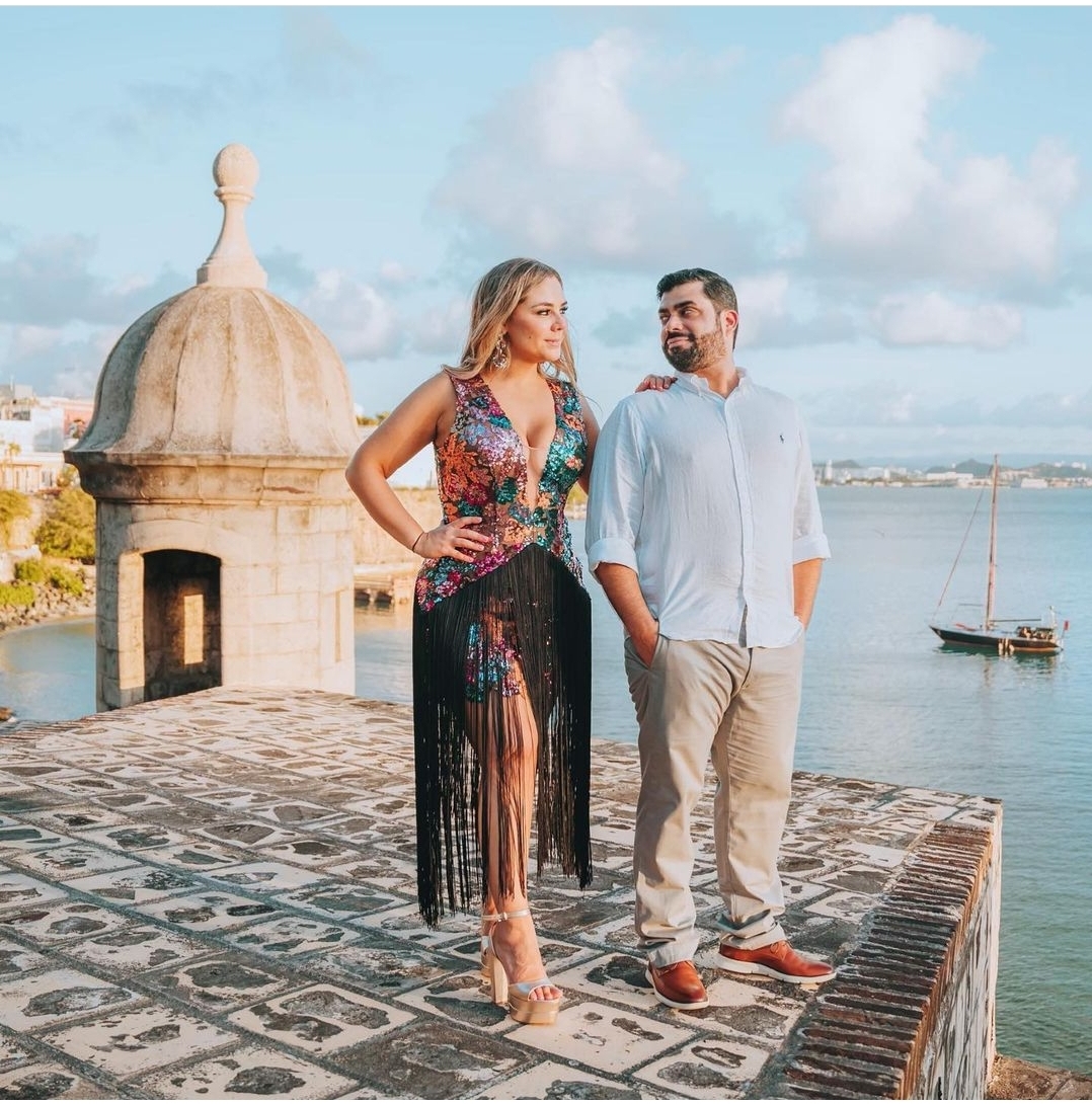 Couple traveling to Puerto Rico experiencing Bucketlist Tours' Old San Juan Ageless Elegance, Heritage and Spirits private tour, on the historic walls of Old San Juan, with a picturesque Guard Tower and stunning views of the bay and sailboat in the backdrop.