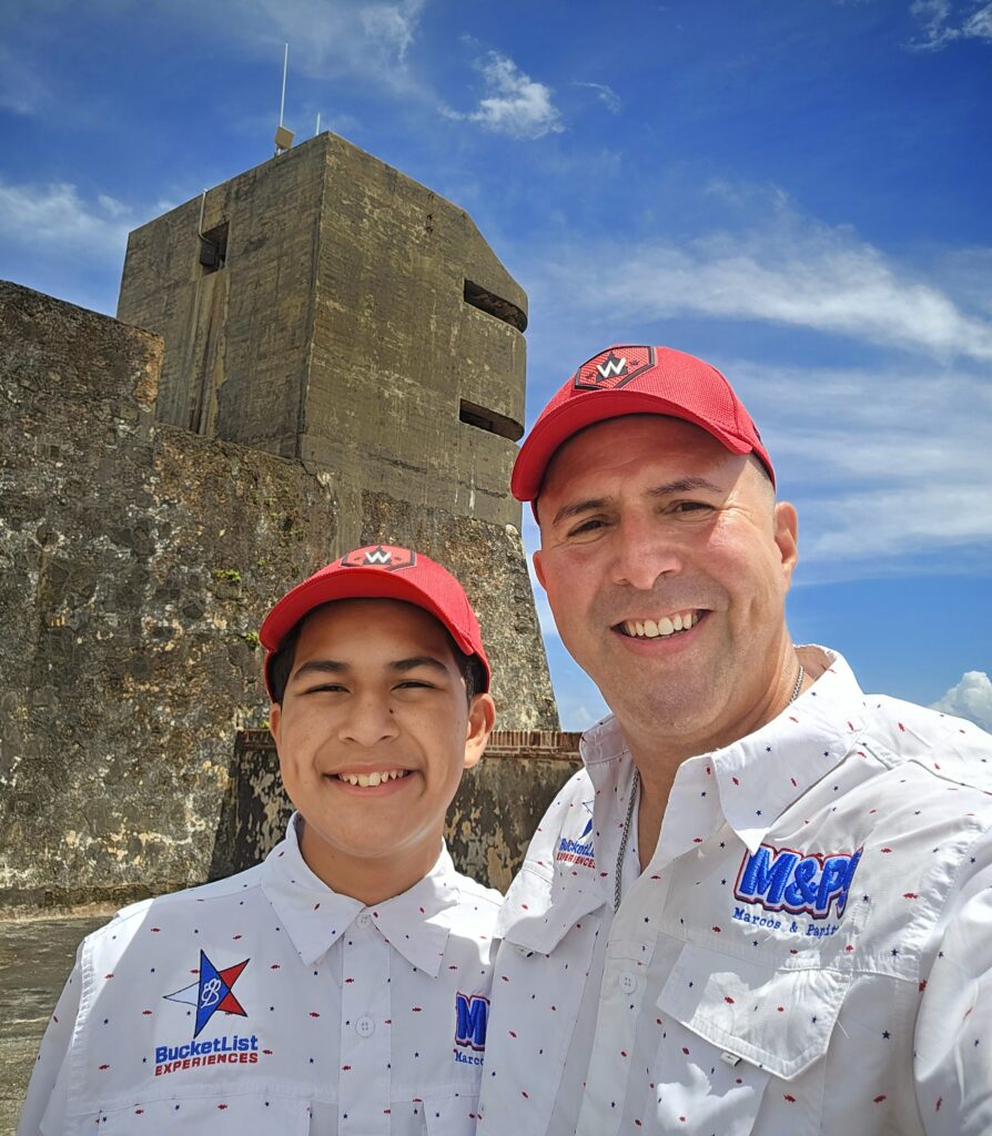 A selfie of Bucketlist Tours Co-Founders, Marcos & Papito!, wearing matching uniforms, like always, on a sunny day in front of a World War II-era tower at Fort San Cristobal in Old San Juan, Puerto Rico. Captured during a Bucketlist Tours Old San Juan's Whispers of Time Private Historic Tour, offered from San Juan, Dorado, and Rio Grande. With their team of passionate locals and travel experts, Marcos & Papito! ensure every tour is infused with unparalleled knowledge, care, and a warm Puerto Rican welcome.