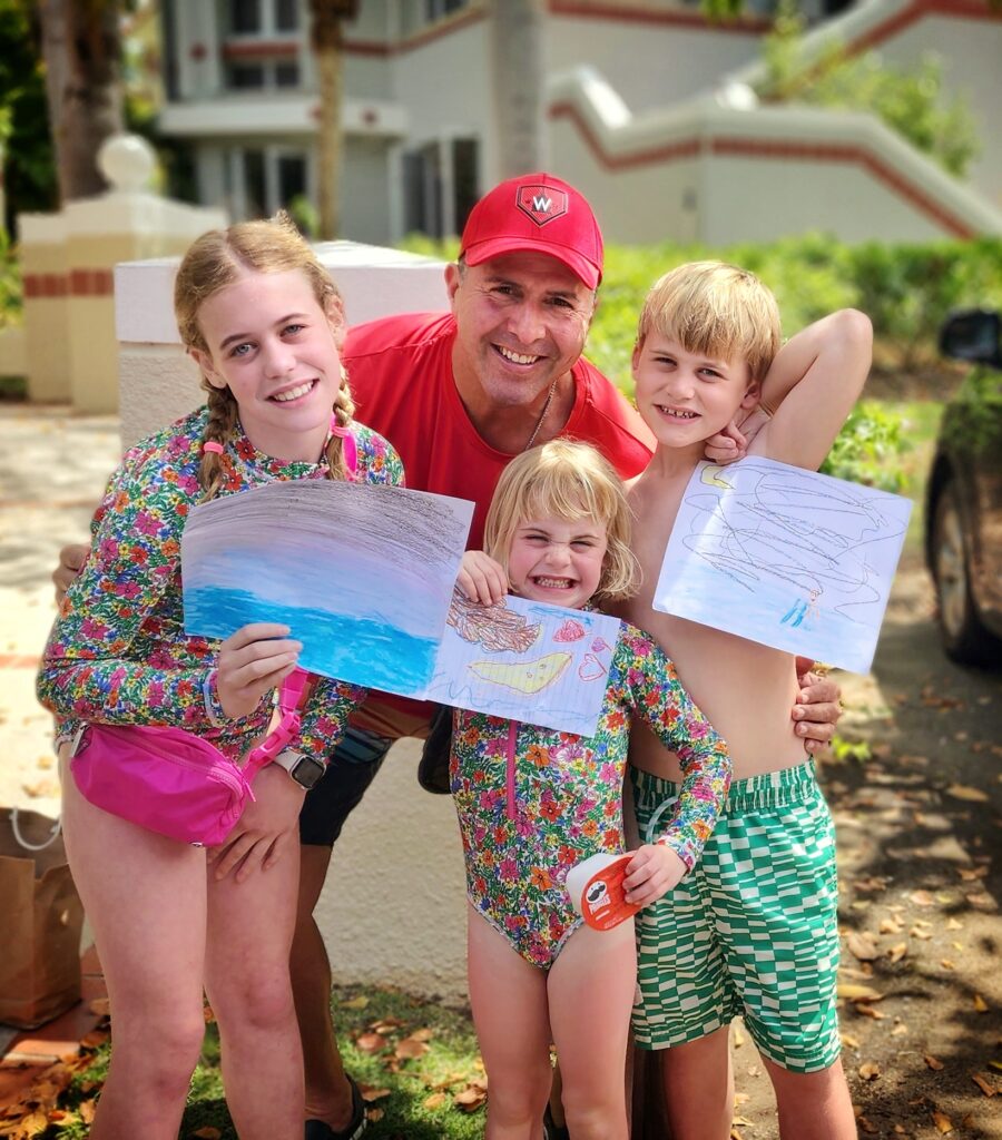 Three children from a family visiting Puerto Rico and stayin in the Ritz Carlton Reserve at Dorado Beach pose with Carlos Alexis, AKA Papito and Bucketlist Tours' Ambassador, holding colorful drawings they made of him swimming in the glowing bioluminescent waters. This heartwarming gesture occurred the morning after the family experienced the Mystic Waters Under the Sun & Sky adventure, part of the Discover Puerto Rico 3-Day Package. These personalized moments highlight the unique and unforgettable experiences Bucketlist Tours delivers.