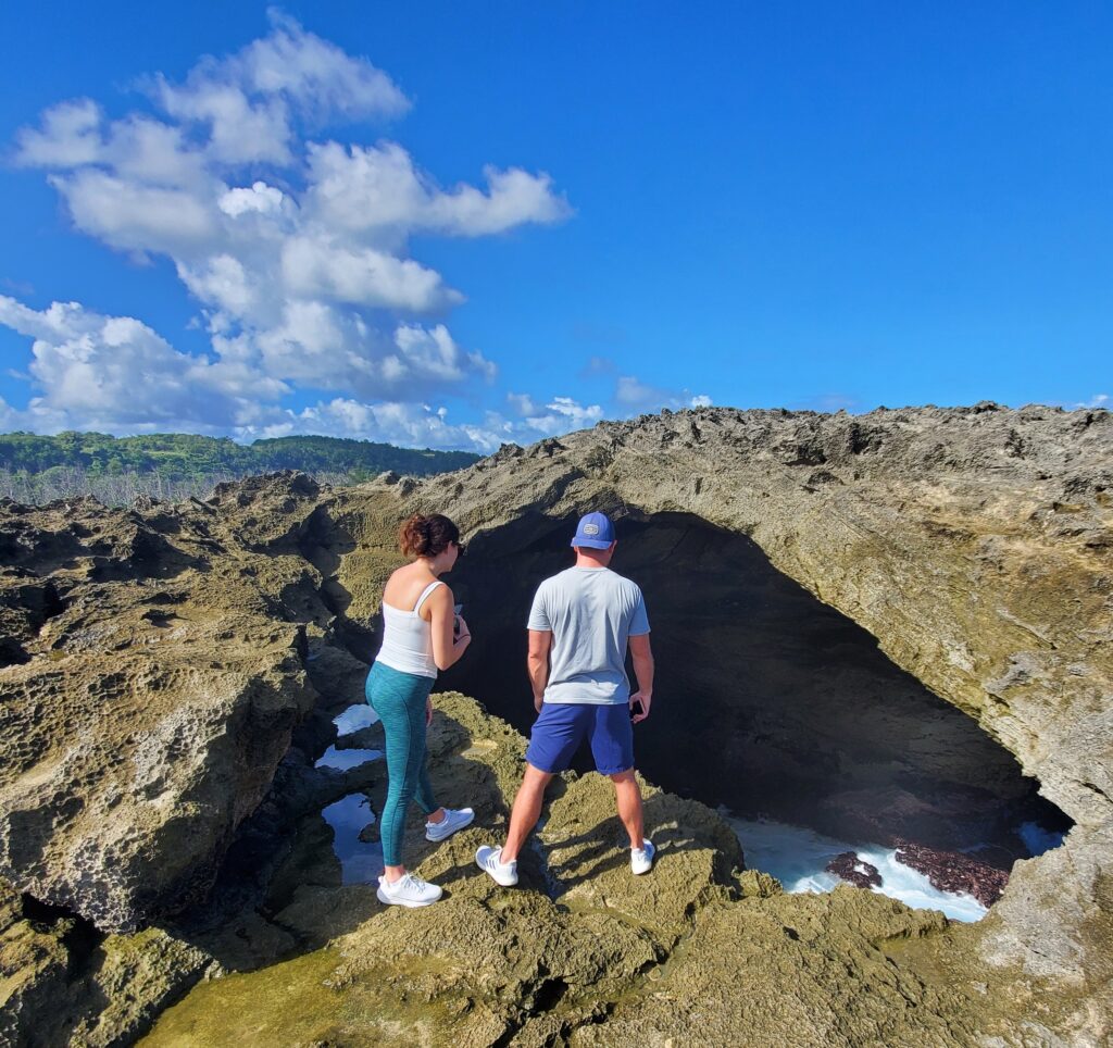 A honeymoon couple stands on the dramatic rock formations of Mar Chiquita, gazing at the ocean waves breaking through a natural blowhole. The scenic view is illuminated by clear blue skies, adding to the magical experience. Their adventure included a safe and moderate hike, allowing them to take in the spectacular coastal landscapes before heading to the Rainforest Waterfalls as part of Bucketlist Tours’ Ocean Edge to Jungle’s Heart private excursion, available from San Juan, Dorado, and Rio Grande.