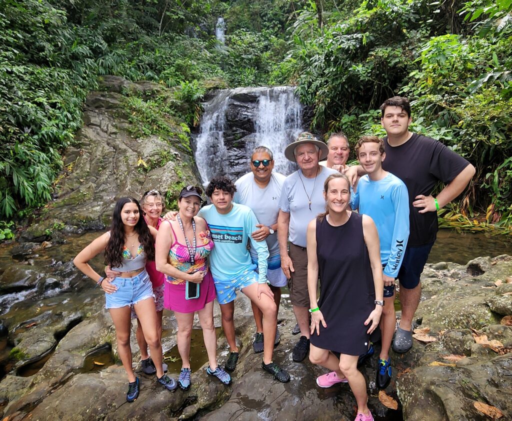 A joyous family spanning three generations smiles as they pose before the serene Las Delicias Waterfalls at Toro Negro. This lively moment captures their anticipation for a cooling swim under the cascading waters, surrounded by lush rainforest scenery. This stop is part of Bucketlist Tours’ Ocean Edge to Jungle’s Heart private adventure, a 6-hour experience starting from San Juan, Dorado, and Rio Grande.