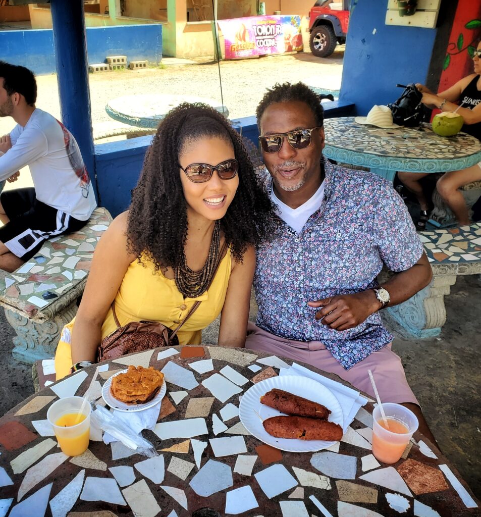 An African American couple savors a delicious meal of Puerto Rico's African-Taino street food fusion while seated at a mosaic-tiled table. The cheerful atmosphere, cultural exploration, and local culinary delights are part of their enriching experience with Bucketlist Tours’ Colonial Spaniard Splendor and African Spirit Private Old San Juan Tour.