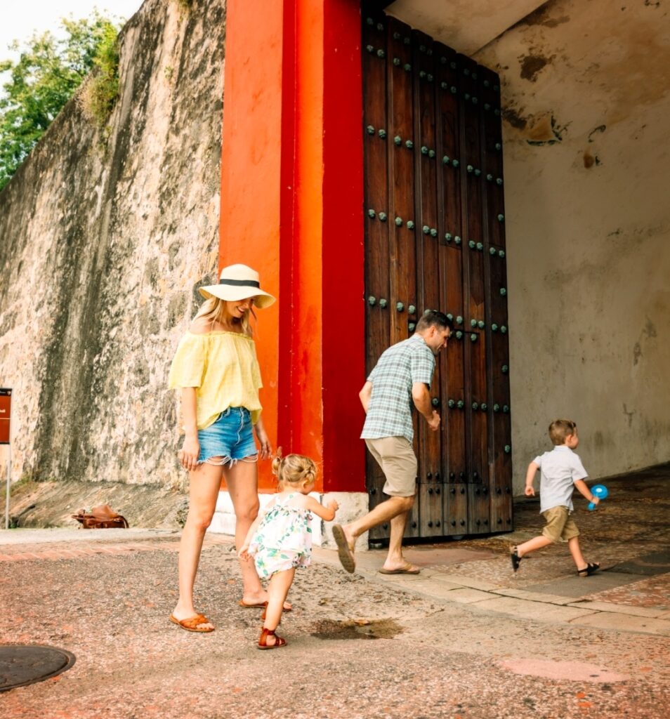 A family with a toddler, strolls through the Gate of San Juan, the entrance to the oldest city center in the Americas. The parents and kids are relaxed and engaged, enjoying the sunny weather and the charm of Puerto Rico’s historic architecture. The vibrant red and wooden gate frames the family, capturing a moment of exploration during Bucketlist Tours’ Colonial Spaniard Splendor and African Spirit Private Old San Juan Tour.