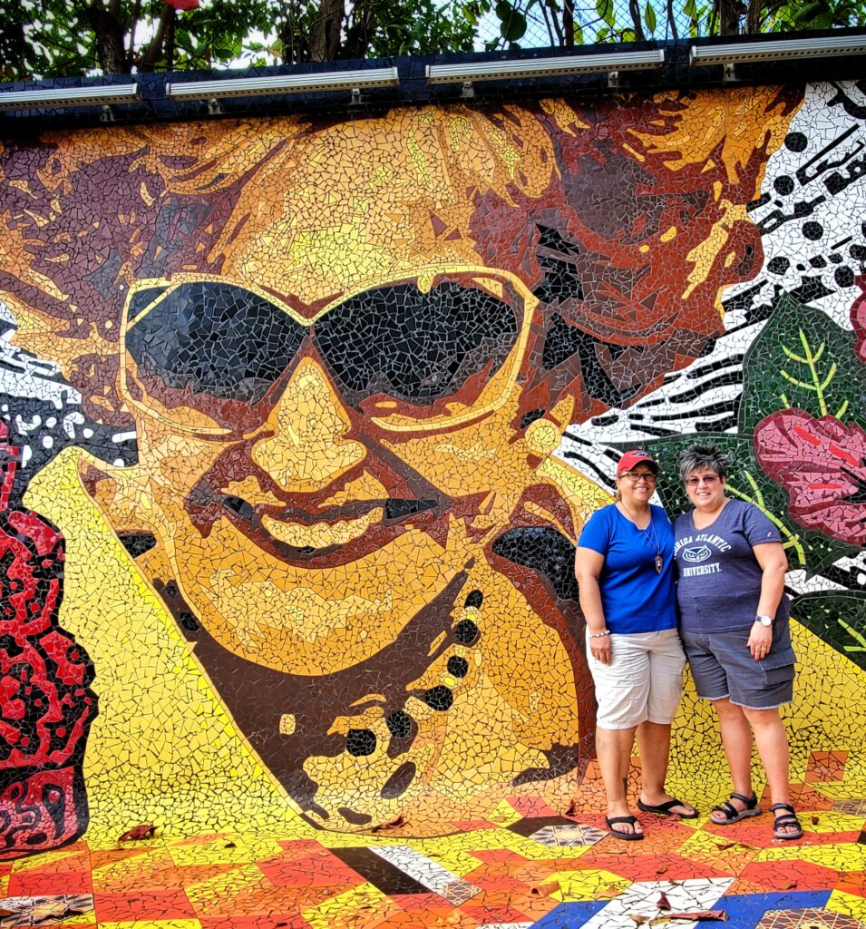 A female couple traveling to Puerto Rico poses in front of Doña Fela’s vibrant mosaic, a memorial honoring Felisa Rincón de Gautier, the first elected female mayor of San Juan. They explore the colorful street art and rich history on a sunny day during Bucketlist Tours’ Colonial Spaniard Splendor and African Spirit Private Old San Juan Tour.