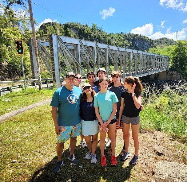 A cheerful family with teens, staying at El Convento Hotel in Old San Juan, poses happily with their guide and ambassador in front of Mata de Plátano Bridge. This historic bridge, built in 1899 by the United States for the train transporting coffee from Ciales to San Juan, marks a significant cultural and historical moment. The stop is part of their Tropical Rainforest Magic & Wonders full-day experience by Bucketlist Tours.