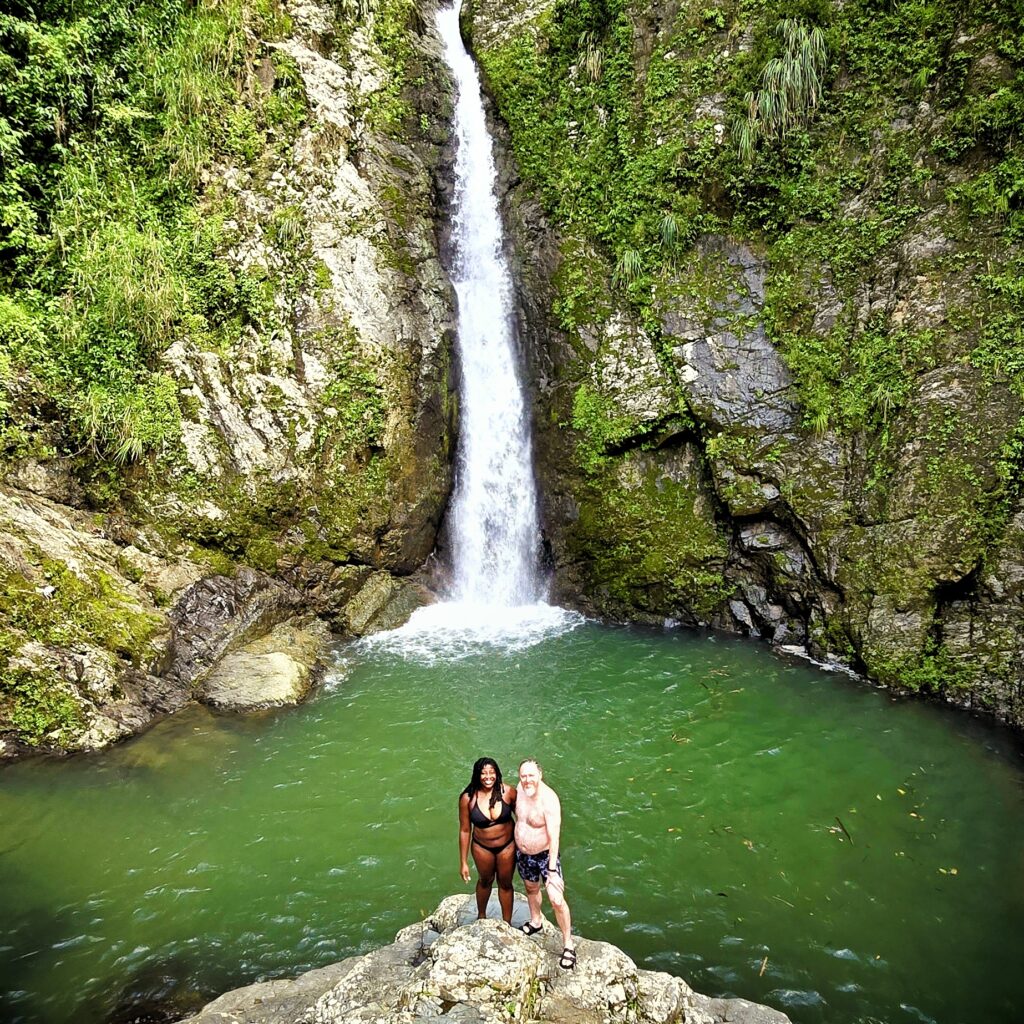 A happy couple staying at El Convento Hotel in Old San Juan poses joyfully in front of Toro Negro's majestic Chorro de Doña Juana Waterfall and its emerald pond. This serene and breathtaking location marks the third stop on their Tropical Rainforest Magic & Wonders full-day experience, an exclusive adventure curated by Bucketlist Tours.