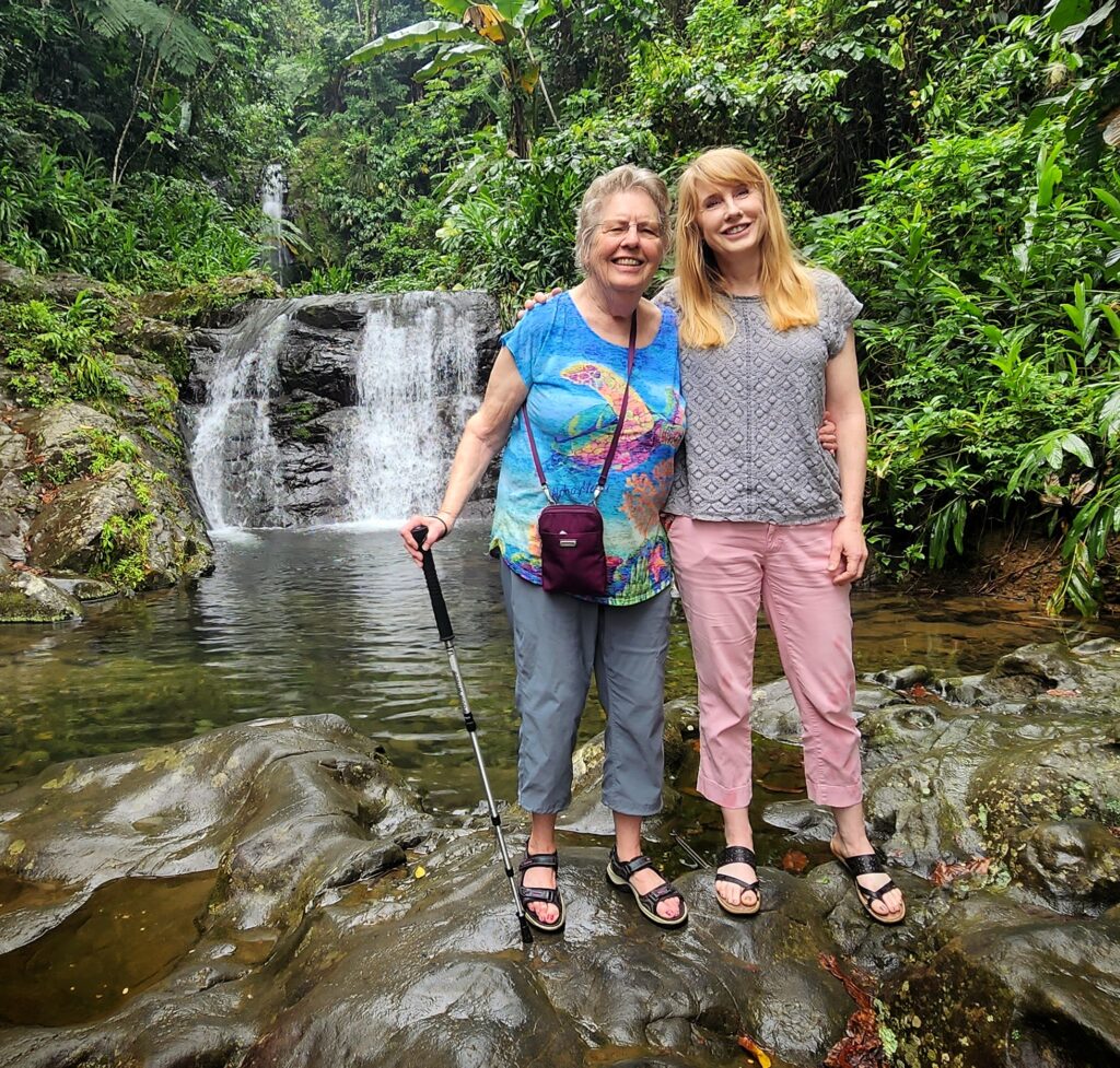 A senior mom and her daughter from Washington State, staying at the elegant El Convento Hotel in Old San Juan, happily pose in front of the serene *Las Delicias waterfall* in *Toro Negro* Rainforest. Captured by their guide and ambassador, this moment celebrates the natural beauty and tranquility of Puerto Rico while they enjoyed their Bucketlist Tours' Tropical Rainforest Magic & Wonders. This experience combine a *roadtrip* with magnificent panoramic views, a *rainforest tour*, a visit to a pond with ancient *Taino petroglyphs* and Puerto Rico's finest *waterfalls*; and was part of their Discover Puerto Rico 3 Day package. The only of its kind, offering an exclusive and unique package delivering the top 10 items of any Puerto Rico Vacation Bucketlist offered offered from San Juan, Dorado, Rio Grande, Isabela and Ponce.