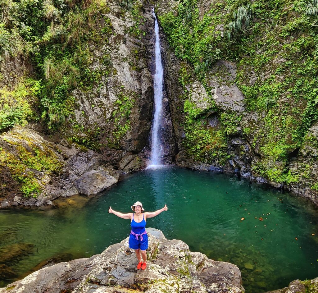 Alma Baird, a frequent VIP guest, and owner at the luxurious Paradise Villas at Ritz Carlton Reserve in Dorado Beach, joyfully poses for her guide and ambassador in front of the majestic Chorro de Doña Juana Waterfall and Pond, the third stop of her Tropical Rainforest Magic & Wonders full-day experience with Bucketlist Tours.