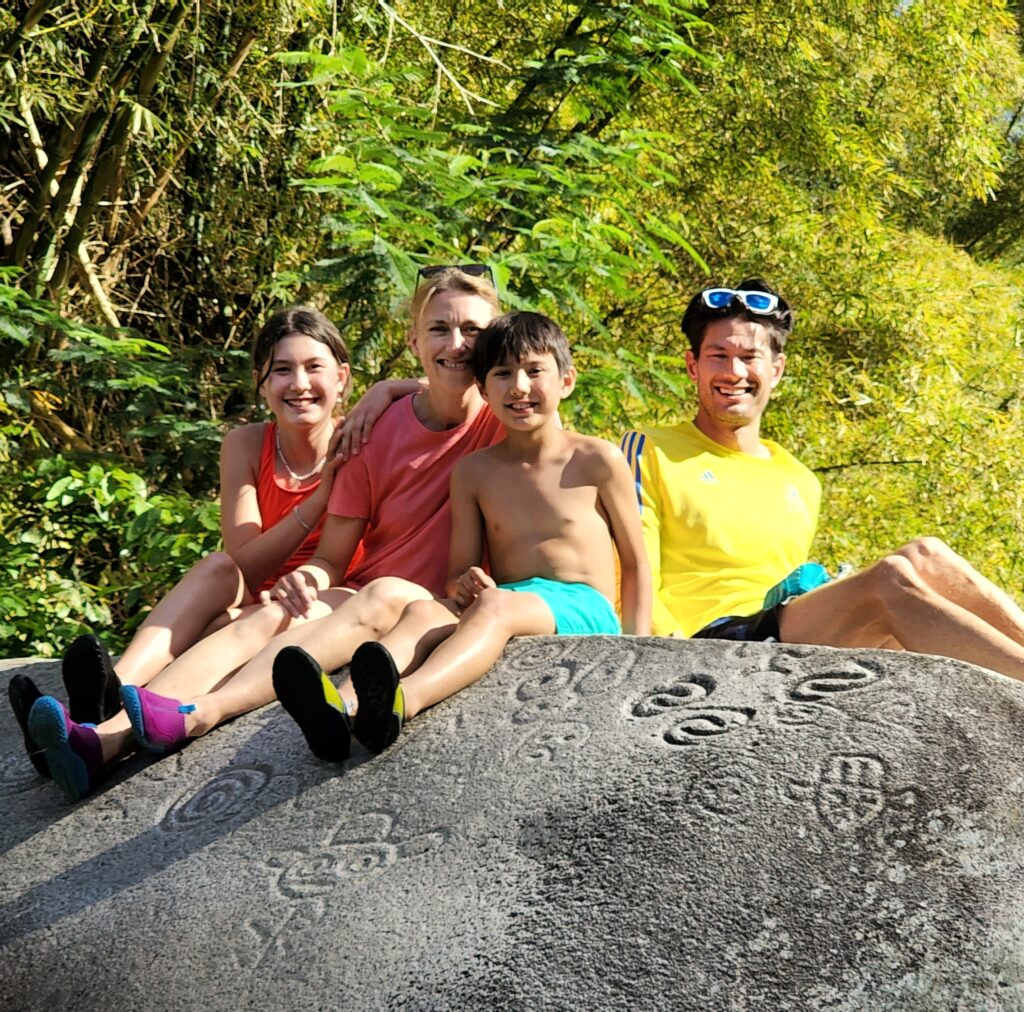 A joyful family of four with children, staying at the Condado Vanderbilt Hotel, poses for their guide and ambassador atop Toro Negro's La Piedra Escrita, a monumental granite rock adorned with ancient Taino petroglyphs. This is the fourth stop of their Tropical Rainforest Magic & Wonders full-day experience by Bucketlist Tours.