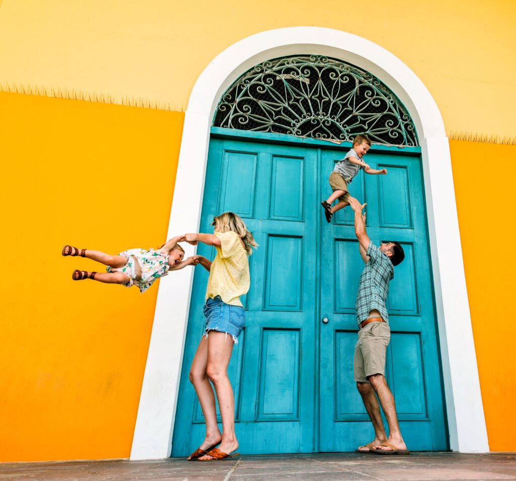 A family of four with young children, traveling to *Puerto Rico* and staying at the Fairmont El San Juan Hotel, is captured joyfully exploring the vibrant architecture of *Old San Juan*. The parents lift their children in playful delight against a backdrop of a striking yellow wall and a turquoise door, showcasing the rich colonial aesthetics of the former farmers’ market. As part of their Timeless Echoes & Legendary Spirits Private Old San Juan Tour, they learn about *Puerto Rico’s evolution* from a *Spanish colony* to a *U.S. territory*, its pivotal role in World War II in defeating Nazi Germany and Japan, and the island's ongoing quest for equal rights and funding parity with the 50 states. This tour combines a *Historic Old San Juan Tour* and a *Rum Distillery Tour*, encompassing over 90 landmarks, including the *Capitol Building*, the *Walkway of the Presidents*, the North Wall, and the iconic streets of the old city. Tailored with safety and comfort in mind, this enriching experience is perfect for couples, families with children, seniors, individuals with mobility constraints, and *cruise ship passengers*.