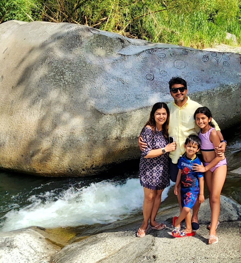 A joyful family with two children from Manhattan, traveling to Puerto Rico and staying in the Ritz Carlton Reserve at Dorado Beach, poses to their guide and ambassador in front of *La Piedra Escrita*, a historic boulder featuring centuries-old Taino petroglyphs, surrounded by the tranquil pond and lush greenery. This moment was captured as they immersed in Puerto Rico's rich cultural and natural heritage on beautiful sunny day as part personalized Bucketlist Tours Tropical Rainforest Magic & Wonders. This experience combine a *roadtrip* with magnificent panoramic views, a *rainforest tour*, a visit to a pond with ancient *Taino petroglyphs* and *Puerto Rico's finest* *waterfalls*; and was part of their Discover Puerto Rico 3 Day package. The only in its kind, offering an exclusive and unique package delivering the top 10 items of any Puerto Rico Vacation Bucketlist offered offered from San Juan, Dorado, Rio Grande, Isabela and Ponce.