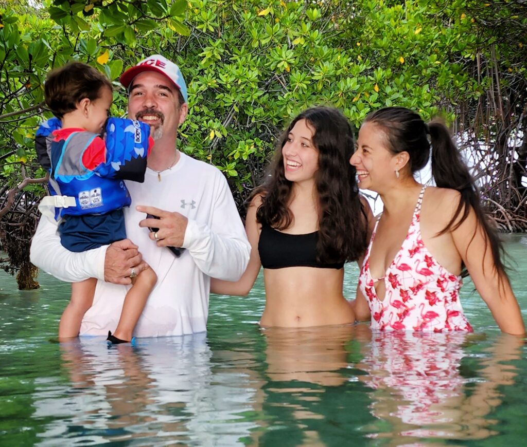 A family, including young children, smiles while wading in the pristine waters of La Parguera's Enrique Cay. Surrounded by lush mangroves, this safe and serene experience is part of Bucketlist Tours’ Mystic Waters Under the Sun & Stars, offering a perfect blend of natural beauty and relaxation for all ages.