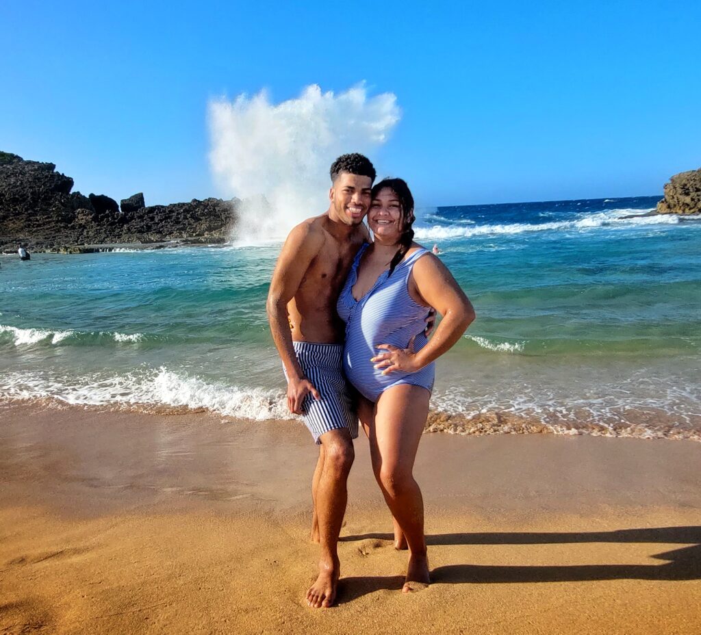 A young Latino couple from Amelia Island beams with joy as they pose for a photo at Poza del Obispo Beach in Puerto Rico. Behind them, an impressive wave splash adds a dramatic backdrop to the stunning sunny day. Staying at the luxurious Condado Vanderbilt, the couple enjoyed this magical moment during Bucketlist Tours' Perfect Beach Escapade: Northern Coastal Adventure, an extension of their Discover Puerto Rico 3-Day Package Private Experience.