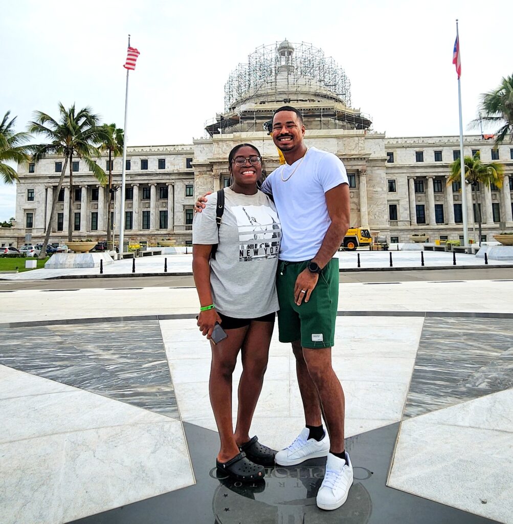 A young African American couple, traveling aboard the *Ritz Carlton Cruise* Ship *Evrima*, poses in front of *Puerto Rico's State Capitol* during their private Old San Juan's Whispers of Time tour. Captured by their guide and ambassador, the couple learns about Puerto Rico's transition from a territory to a commonwealth, the granting of U.S. citizenship to Puerto Ricans, and their ongoing quest for equal rights and parity in government funding with the 50 states. The comprehensive tour explores the *San Juan islet*, covering over 90 landmarks, including the Capitol Building, the *Walkway of the Presidents* (honoring U.S. presidents who visited the island while in office), the northern walls, and the main streets of the historic city. The tour is the perfect *shoreline excursion* for cruiseship passengers being offered in three lengths—2 hours for a comfortable sightseeing tour in a luxury air-conditioned vehicle, 3 hours with 2–3 stops for photos and exploration, or 4 hours with 4 extended stops—the experience is designed with safety, comfort, and accessibility in mind, catering to couples, families, seniors, and individuals with mobility constraints.