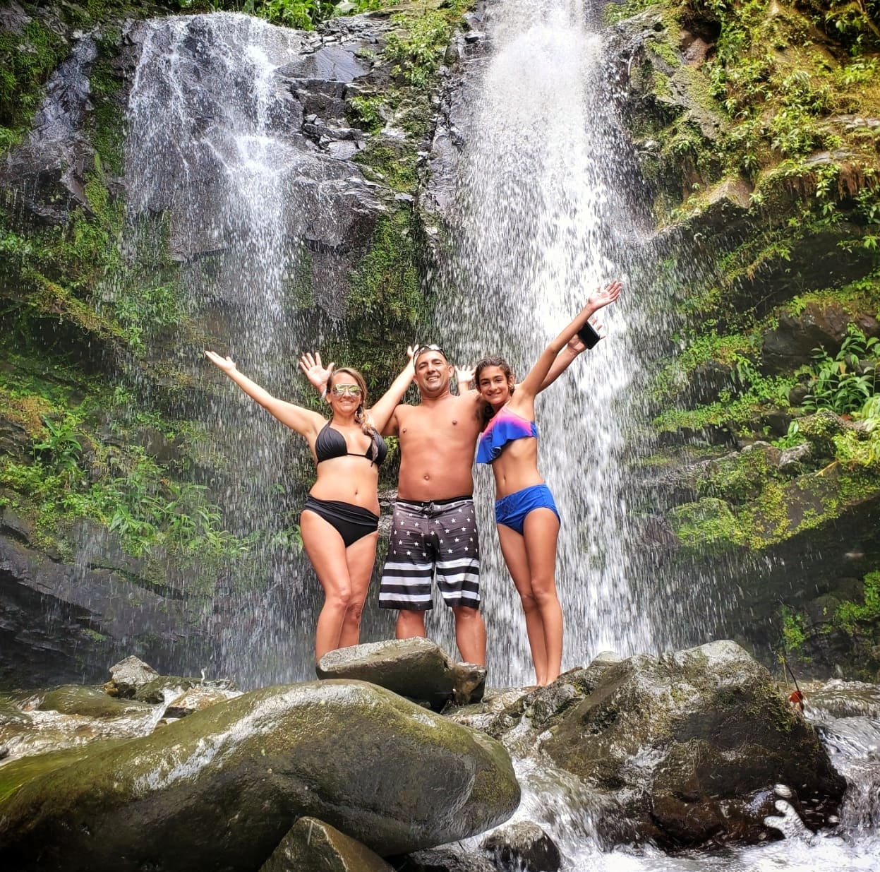 A vibrant family of three, including a teenage girl, staying at the luxurious Condado Vanderbilt Hotel, poses with joy in front of Toro Negro's breathtaking Las Delicias Waterfalls. This moment captures the spirit of their Tropical Rainforest Magic & Wonders full-day experience by Bucketlist Tours, an immersive journey in to the lush beauty of Puerto Rico's natural wonders.